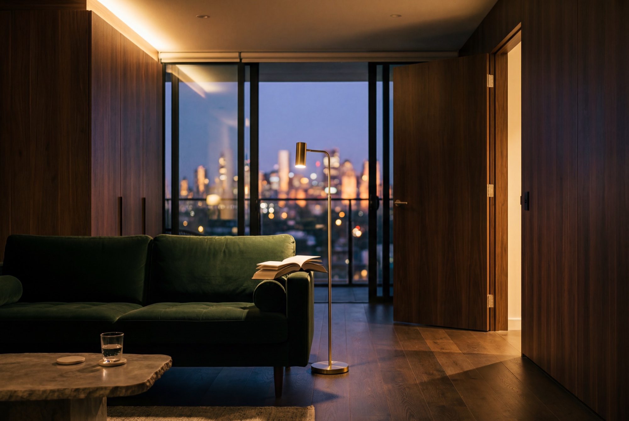A penthouse living room at dusk, cove-lit walnut shelving and a green velvet sofa against the glass