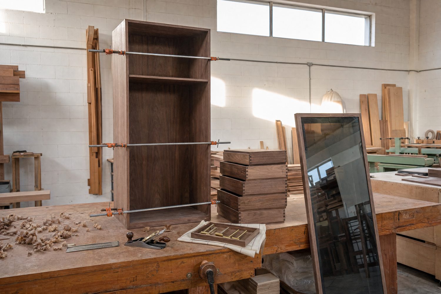 A walnut wardrobe carcass in sash clamps on the Al Quoz bench, dovetailed drawer boxes stacked beside it, a smoked-glass door leaning against the bench