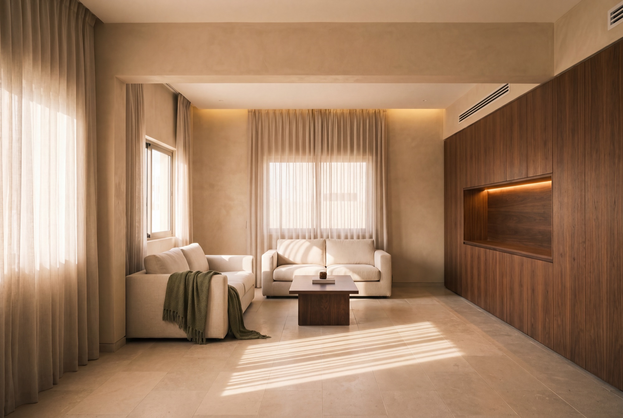 The same room finished – plaster walls, limestone floor, a walnut joinery wall with a cove-lit niche, morning light striping the stone