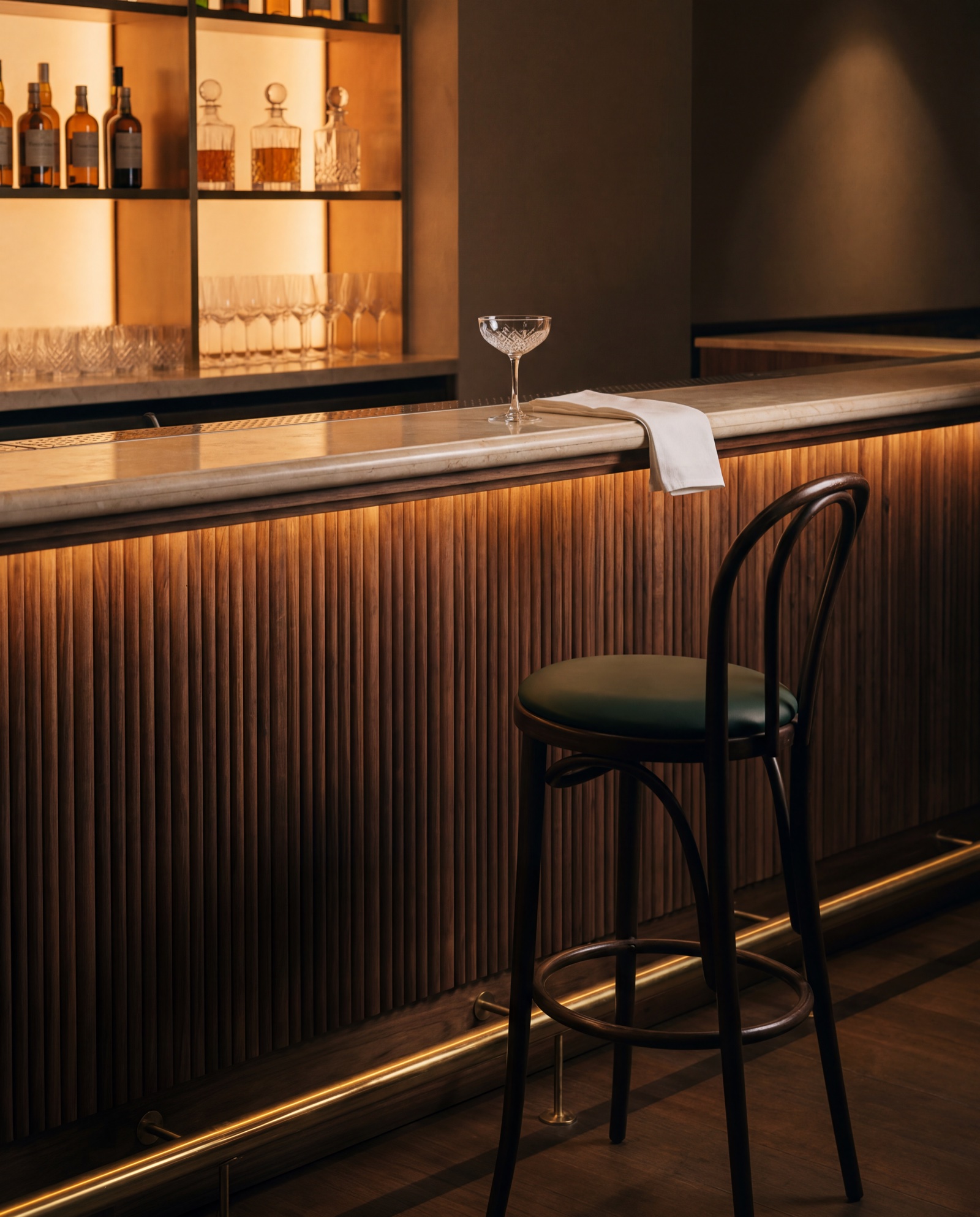 The bar close up – fluted walnut front, brass foot rail catching the light, backlit shelf glowing above