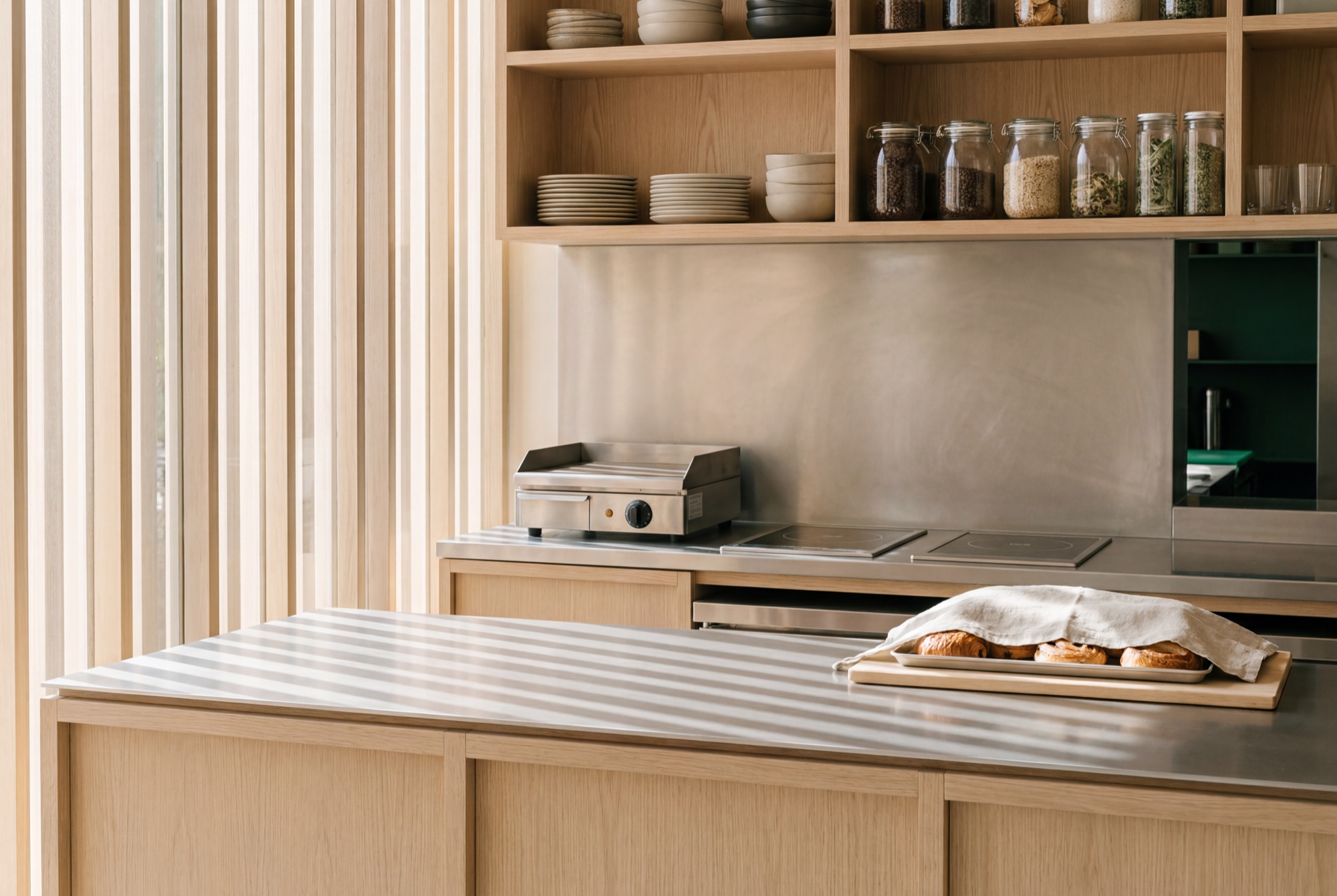 An open café kitchen behind the counter – a short stainless line framed in oak, pastries cooling under a linen cloth