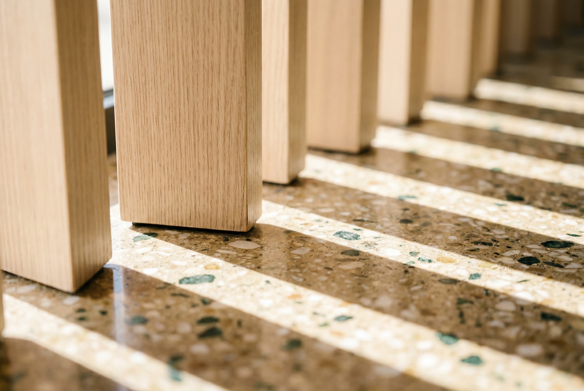 The foot of the oak fin screens meeting the terrazzo floor, morning sun raking through in bars of light