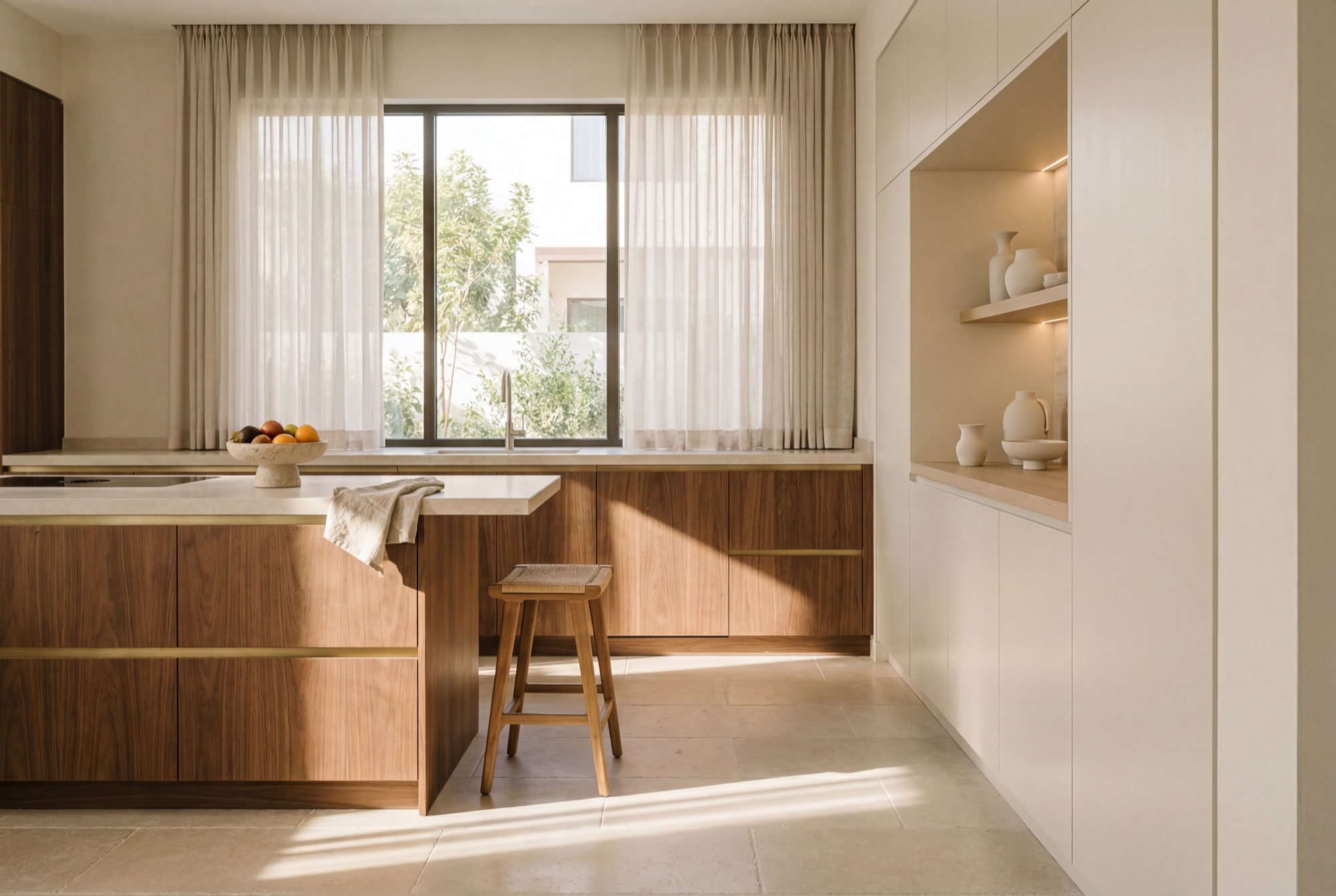 A family kitchen – walnut cabinetry, limestone worktop, morning sun through sheer linen, one stool pulled out